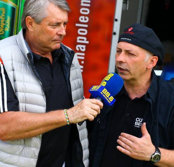 Ein Mann in einem schwarzen Shirt interviewt einen anderen Mann in einer weißen Weste mit einem Mikrofon, auf dem '98.6' steht. Hinter ihnen ist ein roter Banner mit dem Wort 'Napoleon'.