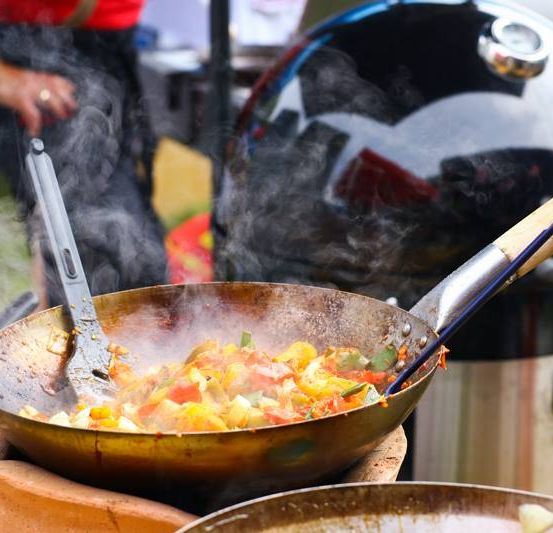 Eine Person kocht Gemüse in einem Wok mit einer Metallzange auf einem Grill. Der Rauch kommt aus dem Grill. Ein Eimer und ein weiterer Wok liegen auf dem Boden.