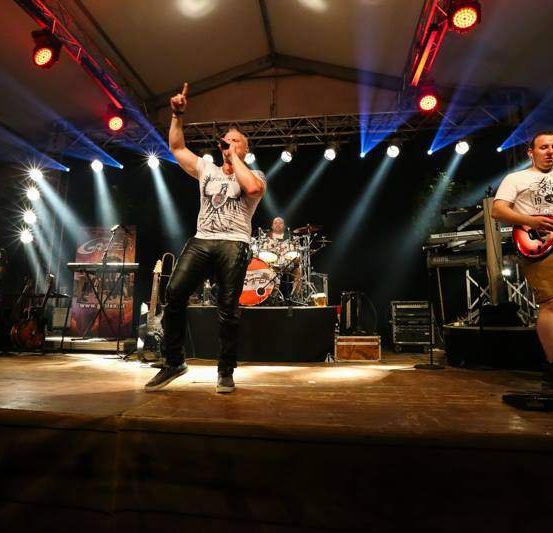 Eine Band tritt auf der Bühne mit Scheinwerfern auf. Der Leadsänger singt und gestikuliert mit der Hand. Der Gitarrist und der Schlagzeuger spielen ihre Instrumente.