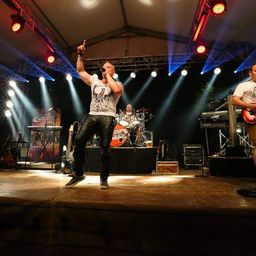 Eine Band tritt auf der Bühne mit Scheinwerfern auf. Der Leadsänger singt und gestikuliert mit der Hand. Der Gitarrist und der Schlagzeuger spielen ihre Instrumente.