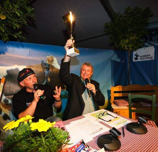 Zwei Männer halten Mikrofone und feiern mit einer Trophäe unter einem Zelt mit Kuhhintergrund. Ein Tisch mit einem rot-weiß karierten Tischtuch hat ein Mikrofonstativ, Blumen und ein Schild.