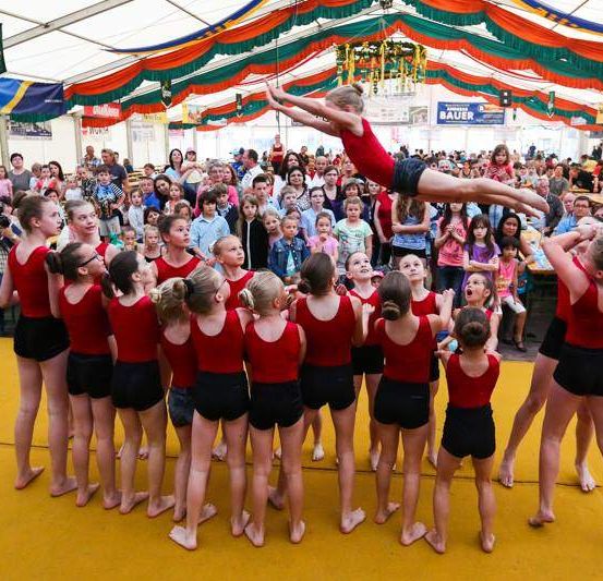 Eine Gruppe von Mädchen in roten und schwarzen Outfits führt eine Tanzroutine vor einem Publikum auf. Eine rot gekleidete Mädchen macht in der Mitte der Gruppe eine Handstand. Das Publikum beobachtet sie.