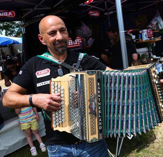 Ein Mann steht im Freien, lächelt und hält ein Akkordeon mit einem farbenfrohen Design. Hinter ihm ist ein Stand mit verschiedenen Artikeln zu sehen. Leute sitzen an Tischen unter Zelten.