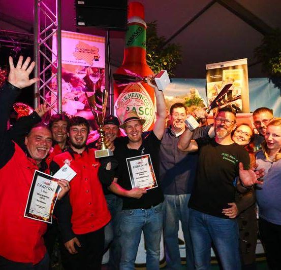 Eine Gruppe von Menschen posiert für ein Foto mit ihren Trophäen und Auszeichnungen bei einer Zeremonie. Der Mann in der Mitte hält eine Flasche Tabasco-Sauce. Hinter ihnen steht ein großes Plakat einer Chilisaucenflasche.