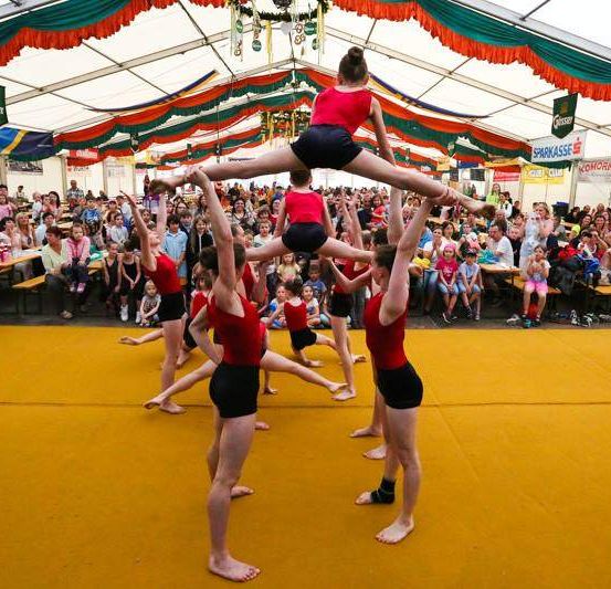 Eine Gruppe von Turnern führt eine akrobatische Routine unter einem Zelt vor, während eine Menschenmenge zuschaut. Eine Turnerin balanciert auf den Schultern anderer.