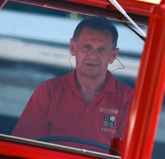 Ein älterer Mann in einem roten Poloshirt mit 'DB' und 'DBTCA' sitzt in einem roten Fahrzeug. Er trägt Kopfhörer und scheint auf die Straße konzentriert zu sein.
