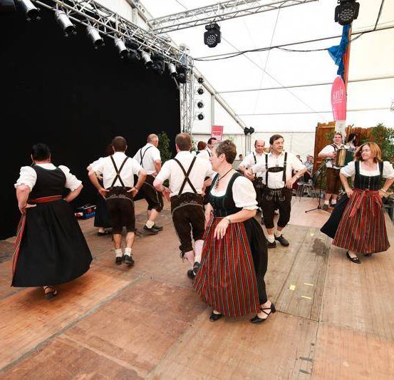 Mehrere Menschen tanzen auf der Bühne in traditioneller bayerischer Kleidung mit schwarzem Hintergrund und Metallkonstruktion darüber.