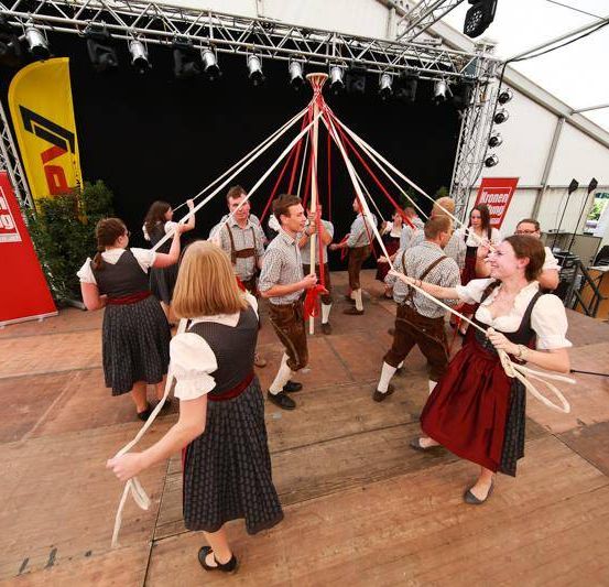 Eine Gruppe von Menschen in traditioneller Kleidung führt einen Tanz mit einem Maibaum auf einer Bühne auf.