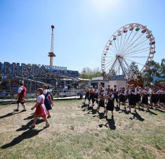 Eine Gruppe von Menschen in traditioneller bayerischer Kleidung geht über ein grasbewachsenes Feld vor einem großen Vergnügungspark. Eine Band spielt Instrumente und ein Riesenrad ist im Hintergrund zu sehen.