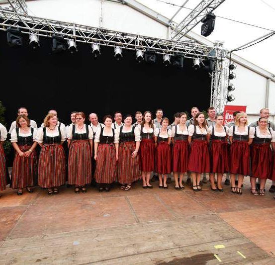 Eine Gruppe von Menschen in traditioneller Kleidung steht auf der Bühne. Hinter ihnen befindet sich ein Hintergrund mit einem Banner. Eine Person in der Ecke hält einen Holzstab.