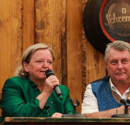 Zwei Personen, eine Frau in Grün und ein Mann in Blau, sitzen an einem Tisch mit Mikrofonen. Hinter ihnen steht eine Holzwand mit einem großen Fass, das 'Schwemser' beschriftet ist, und einem Plakat mit Telefonnummern.