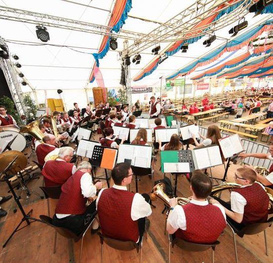 Eine Band spielt in einem großen Saal vor einem großen Publikum, das an Tischen sitzt und von Deckenleuchten beleuchtet wird. Die Bandmitglieder tragen rote Westen und spielen Instrumente wie Trompeten und Posaunen.