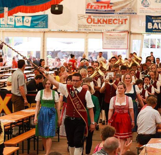Eine Gruppe von Menschen in traditioneller deutscher Kleidung, die mit Blechblasinstrumenten in einem großen Zelt marschiert, möglicherweise bei einem Festival oder einer Veranstaltung.