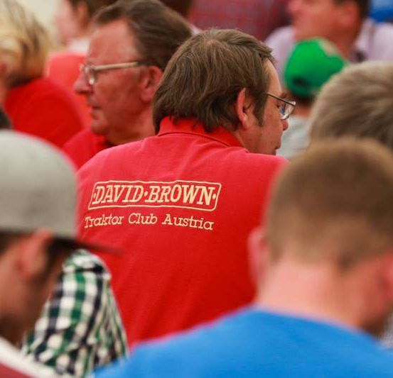 Eine Gruppe von Menschen nimmt an einer Veranstaltung teil. Einige tragen rote T-Shirts mit den Aufschriften 'DAVID BROWN' und 'Traktor Club Austria'. Ein Mann mit Brille schaut sich etwas an.
