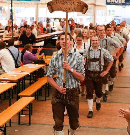 Eine Gruppe von Männern in traditioneller bayerischer Kleidung geht durch ein überfülltes Bierzelt und trägt ein Schild.
