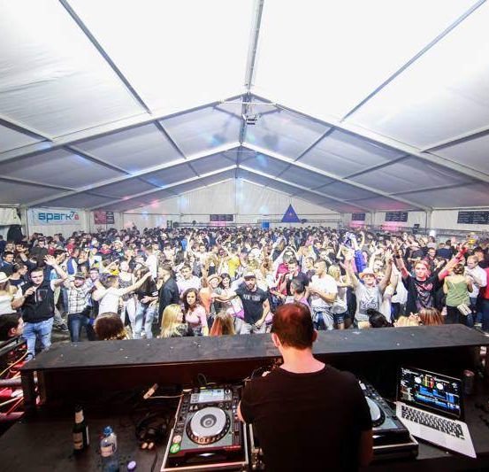 Eine große Menschenmenge tanzt bei einer Musikveranstaltung unter einem Zelt, vor einem DJ mit DJ-Equipment.