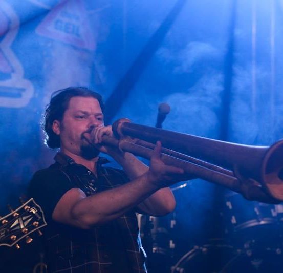 Ein Mann spielt auf einem Didgeridoo unter einem blauen Bühnenlicht mit einer Gitarre und Trommeln im Hintergrund.