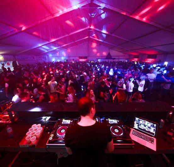 Ein DJ spielt Musik in einem überfüllten Nachtclub mit roter und blauer Beleuchtung. Viele Leute tanzen vor der DJ-Konsole.