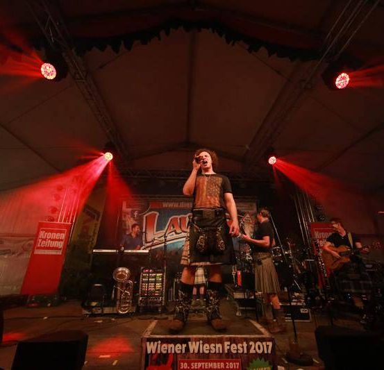 Eine Band tritt auf einer Bühne unter roten Lichtern auf. Ein Mann singt, während er auf einem Kasten steht. Hinter ihm spielen andere Bandmitglieder verschiedene Instrumente.