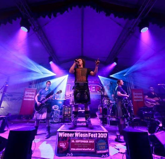 Eine Band tritt auf der Bühne mit lila Beleuchtung beim Wiener Wiesn Fest 2017 auf. Ein Leadsänger steht auf einer Plattform, während andere Instrumente spielen.