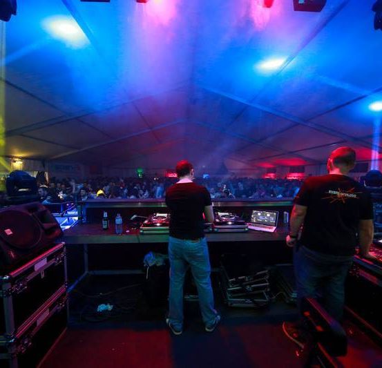 Zwei DJs spielen Musik in einem hell beleuchteten Zelt. Einer steht neben DJ-Ausrüstung, während der andere zuschaut. Ein Publikum füllt den Raum.
