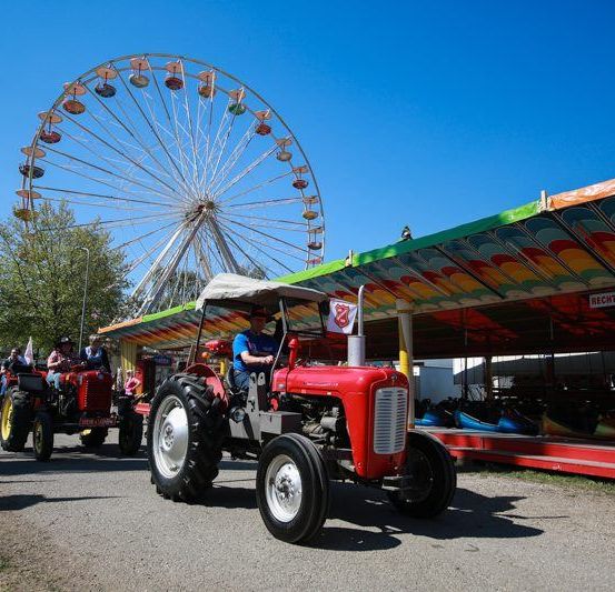 Eine Gruppe Traktoren fährt an einem bunten Jahrmarkt vorbei, mit einem großen Riesenrad und verschiedenen Attraktionen im Hintergrund.