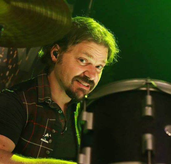 Ein Mann mit grünen Haaren spielt lächelnd Schlagzeug unter grünem Licht.