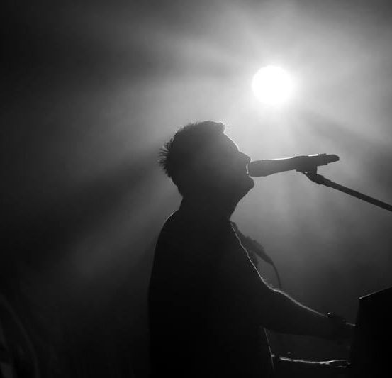 Die Silhouette eines Mannes spielt Klavier und singt in ein Mikrofon, beleuchtet von einem hellen Licht.