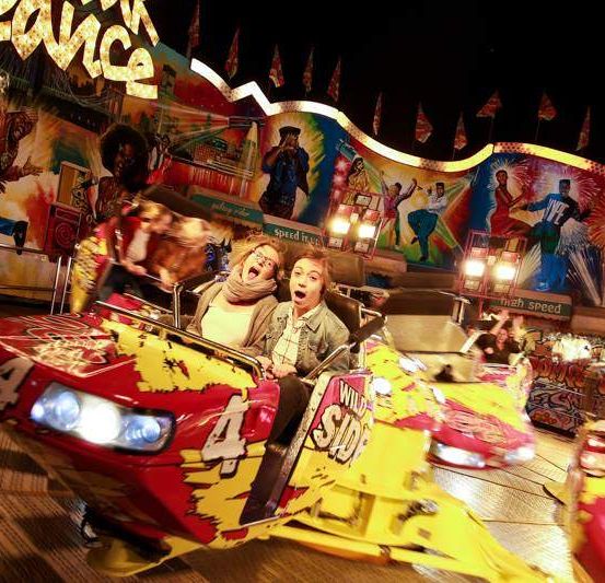 Zwei Personen fahren bei Nacht eine farbenfrohe Achterbahn im Jahrmarkt. Die Achterbahn ist rot und gelb bemalt mit einer Nummer vier auf der Vorderseite. Sie sind von hellen Lichtern und bunten Dekorationen umgeben.