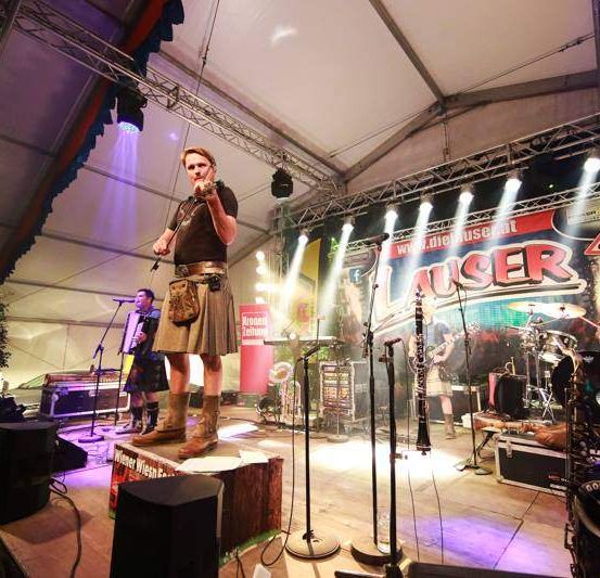 Ein Mann spielt Geige auf der Bühne mit einer Band, Scheinwerfer beleuchten die Aufführung und ein Publikum ist anwesend.