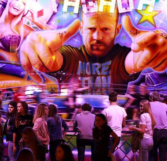 Eine große Menschenmenge versammelt sich auf einem Jahrmarkt und betrachtet ein großes Wandbild eines Mannes mit den Worten 'Mehr als'. Das Wandbild ist farbenfroh und enthält Bilder von Autos und Frauen.