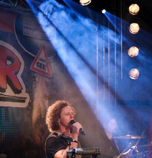 Ein Mann singt auf der Bühne mit blauen Scheinwerfern und Nebel. Dahinter spielt ein Schlagzeuger Trommeln und Becken. Der Hintergrund zeigt ein rotes und weißes Schild mit 'R' und einem Warndreieck.