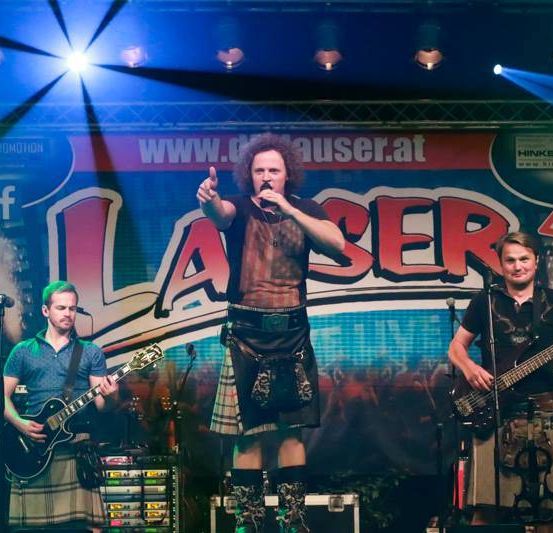 Eine Band tritt auf der Bühne auf, wobei ein Scheinwerfer auf den Leadsänger gerichtet ist. Ein Mann spielt Gitarre und ein anderer Schlagzeug. Ein Banner mit dem Wort 'Laser' ist hinter ihnen angebracht.