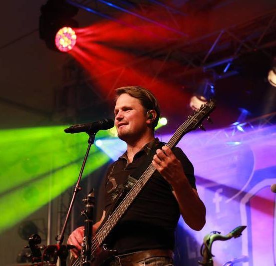 Ein Mann spielt Gitarre auf der Bühne mit einem Mikrofon vor ihm. Er schaut nach rechts. Hinter ihm steht ein Saxophon. Die Bühne wird von grünen und roten Lichtern beleuchtet.