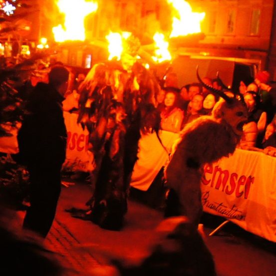 Eine Nachtszene zeigt eine Menge, die einen Ritus beobachtet. Zwei Figuren in Tierkostümen halten Fackeln. Eine hat Hörner, die andere trägt eine Maske. Sie stehen vor einem Banner.