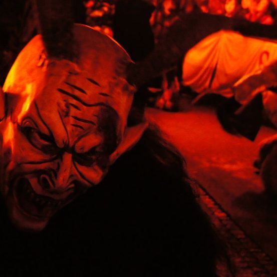 Eine Person mit einer dämonischen Maske mit leuchtenden Augen und scharfen Zähnen ist in einem schwach beleuchteten Raum zu sehen. Eine Person liegt im Hintergrund auf dem Boden.