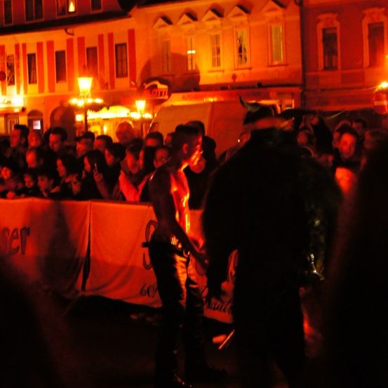 Eine Menschenmenge versammelt sich draußen und sieht eine Aufführung. Ein Mann mit Hörnern steht davor, während eine andere Person ein Schwert hält. Banner sind an einer Absperrung befestigt. Gebäude mit beleuchteten Fenstern sind im Hintergrund.