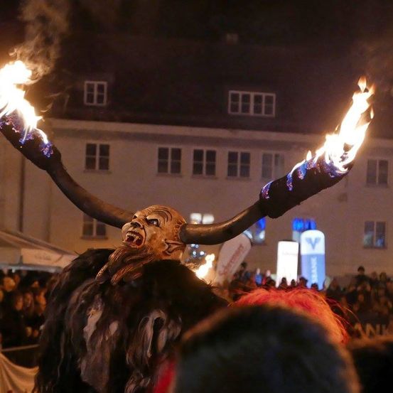 Eine Person in einem gehörnten Kostüm mit brennenden Fackeln steht bei Nacht vor einer Menschenmenge, mit Gebäuden und Lichtern im Hintergrund.