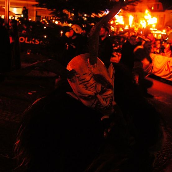 Eine Person in einer Dämonenmaske steht in einer Menschenmenge. Dahinter sehen Menschen ein Feuer. Im Hintergrund steht ein Gebäude mit einer Weihnachtsbaum und Lichtern.