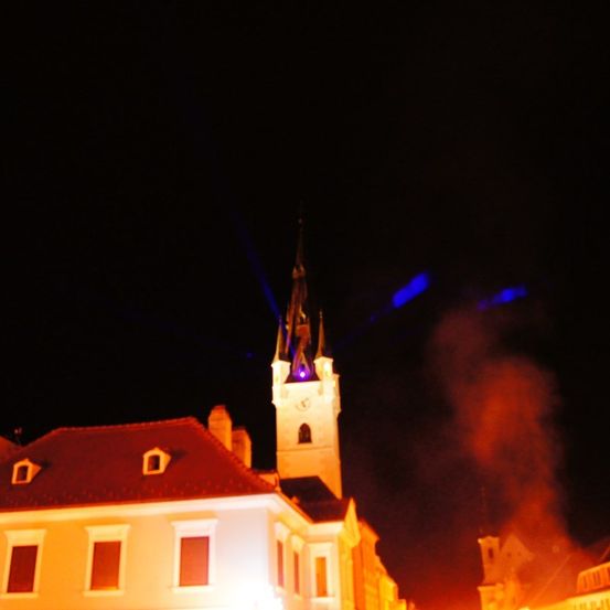 Ein Gebäude mit einem roten Dach und mehreren Fenstern wird nachts von einem blauen Licht beleuchtet. Das Licht kommt von der Spitze eines hohen Turms.