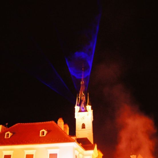 Eine Nachtszene mit einer Kirche, bei der ein blaues Laserlicht in den Himmel strahlt und Rauch aus dem Schornstein aufsteigt.
