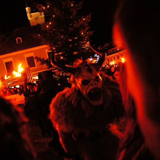 Eine Person in einem Hörnerkostüm steht bei Nacht vor einem Gebäude mit einem beleuchteten Baum und einem Feuer inmitten einer Menschenmenge.