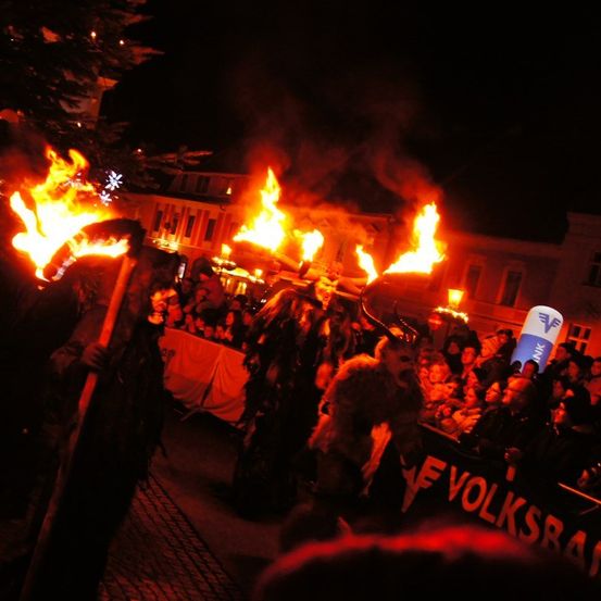Eine Gruppe von Menschen in Kostümen und Masken geht bei Nacht durch eine Menschenmenge und hält brennende Fackeln in den Händen.
