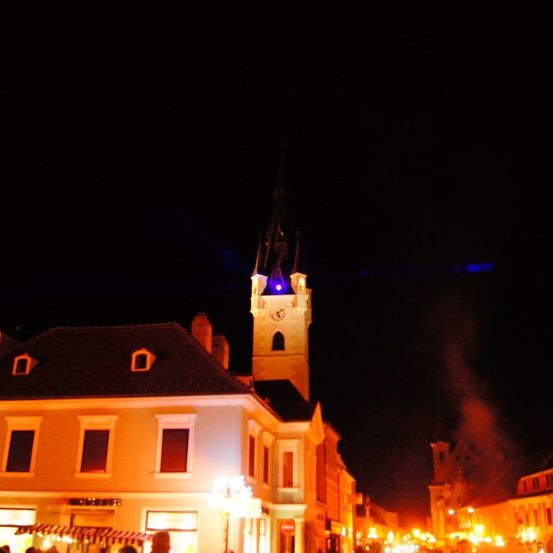 Eine Nachtszene einer Stadt mit Gebäuden, einem Turm mit einer Uhr und Lichtern sowie Menschen, die auf der Straße gehen.