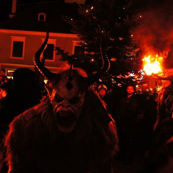 Eine Person in einem gehörnten Kostüm tritt bei einem nächtlichen Festival auf, mit einem brennenden Baum im Hintergrund.