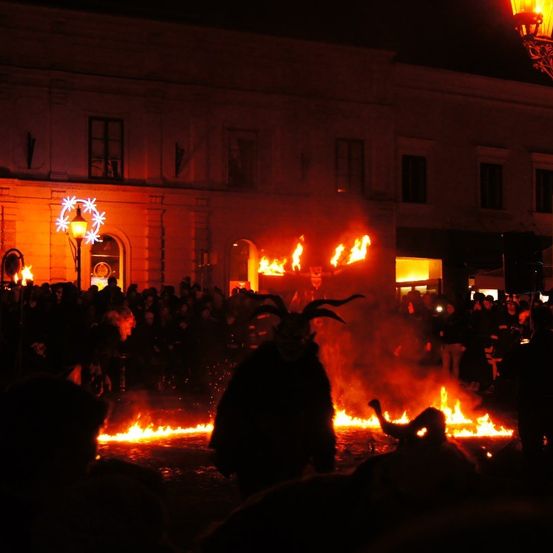 In der Nacht versammelt sich eine Menge um ein großes Lagerfeuer, einige tragen Hörner. Das Feuer brennt hell und wirft ein warmes Licht. Ein Gebäude mit vielen Fenstern steht im Hintergrund.