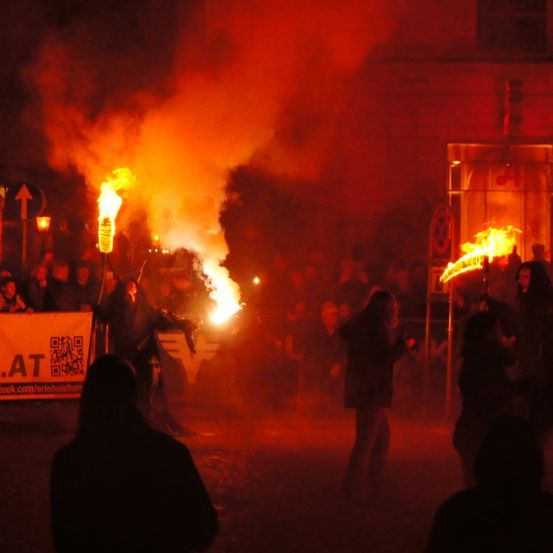 Eine nächtliche Versammlung mit Menschen, die brennende Fackeln halten und vor einem Gebäude mit rotem Schein stehen. Ein weißes Banner mit Text und einem QR-Code ist sichtbar.