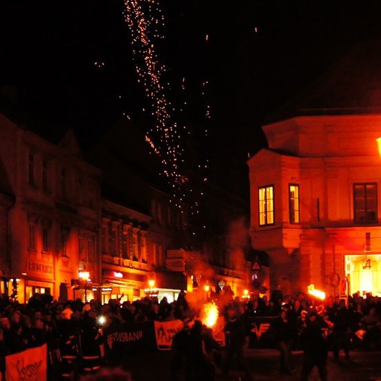 Eine große Menschenmenge versammelt sich nachts auf einem Stadtplatz, hält Transparente und Fackeln. Gebäude umgeben sie, und Feuerwerke erleuchten den Himmel.