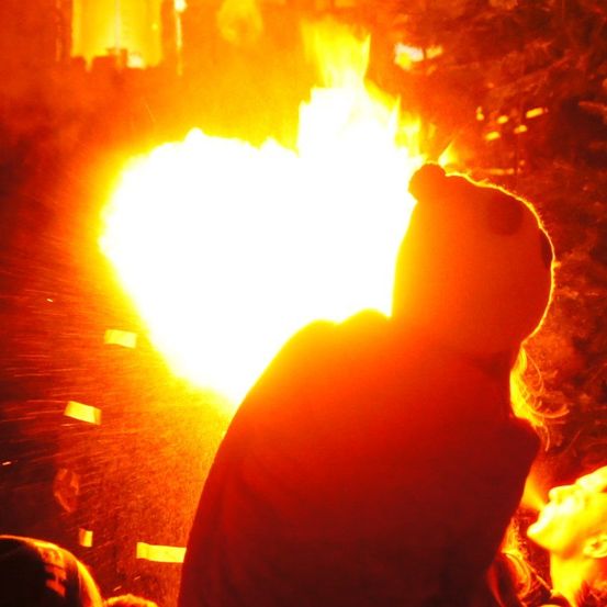 Eine Person mit einem Panda-Hut beobachtet ein Feuer, das von Rauch umgeben ist. Eine weitere Person steht in der Nähe.