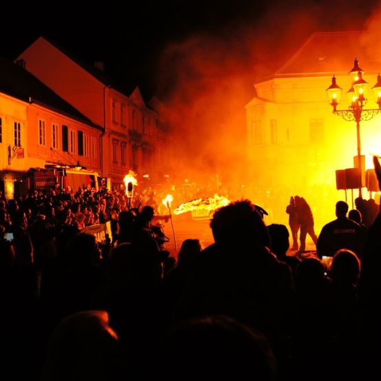 Eine nächtliche Straßenszene mit einem brennenden Fahrzeug im Vordergrund. Menschenmengen versammeln sich darum, einige filmen mit Handys. Ein Feuerwehrwagen ist in der Nähe geparkt.